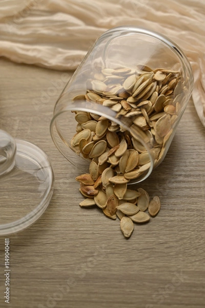 Obraz Scattered pumpkin seeds, in transparent glass jar on wooden table, beige cloth on the background