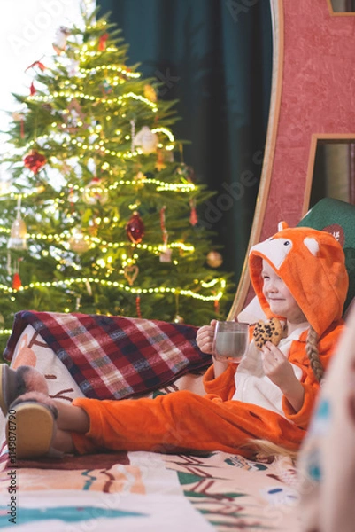Obraz Christmas, New Year's photo. A child girl in a fox cub costume is sitting on the couch, smiling, drinking milk and eating cookies. There is a Christmas tree in the background.