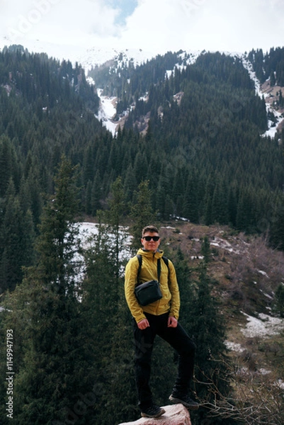 Obraz A young man in sunglasses stands against the backdrop of snow-capped mountain peaks. Trekking in the mountains