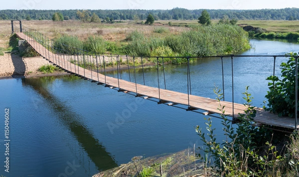 Fototapeta Unusual design. Suspended wooden bridge on steel cables or ropes. Not a wide river. Summer day. Forest, bushes, water, shadow and reflection of the bridge in the water.