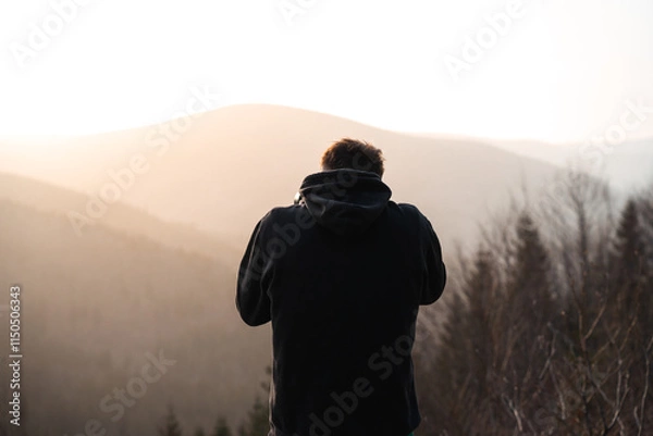Fototapeta A man dressed in a black hoodie is standing in front of a vast mountain range