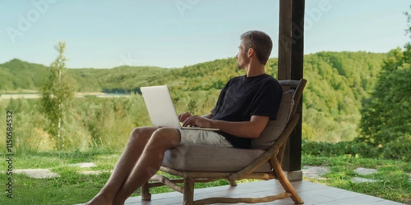 Obraz Young male freelancer seated on a wooden chair, with laptop, enjoying nature and a beautiful forest view while working remotely. Work-life balance. Copy space
