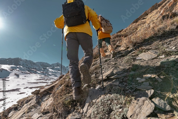 Fototapeta Travelers and friends climb the mountain with sticks and backpacks against the background of mountains and sky, view from the back from the bottom. People are walking with backpacks in bright clothes.