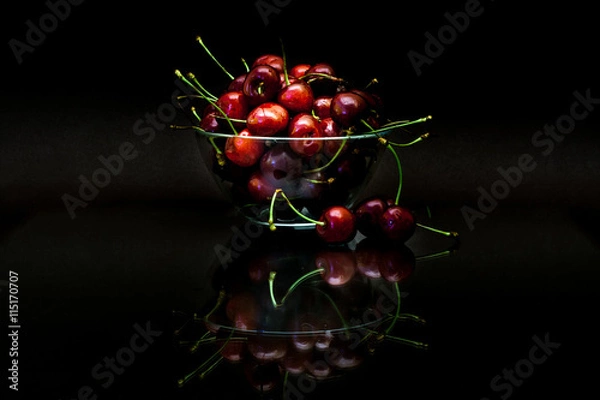 Fototapeta Transparent cup with red ripe cherries with reflection on black background.