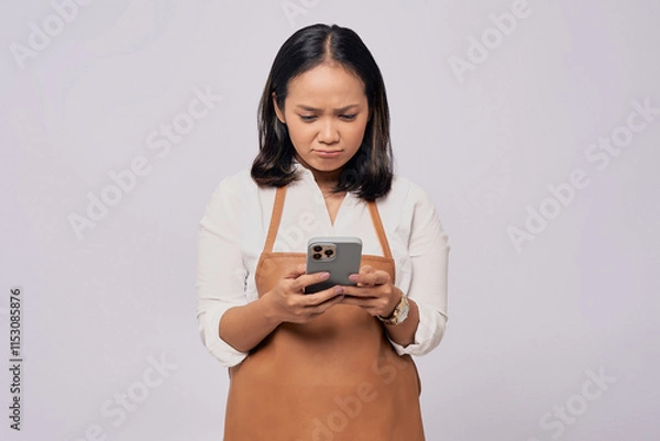 Fototapeta Unhappy young Asian woman barista barman employee wearing brown apron working in coffee shop using mobile phone isolated on white background. Small business startup concept