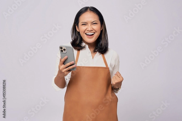 Fototapeta Smiling young Asian woman barista barman employee wearing a brown apron working in coffee shop holding mobile phone and celebrating success isolated on white background. Small business startup concept