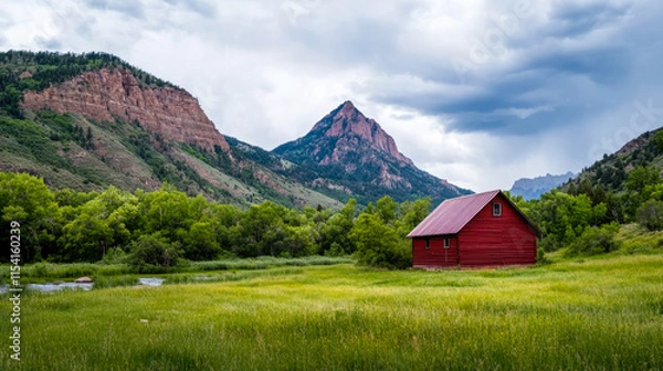 Fototapeta Serene red barn nestled in a lush valley surrounded by stunning mountains in Colorado during a cloudy afternoon