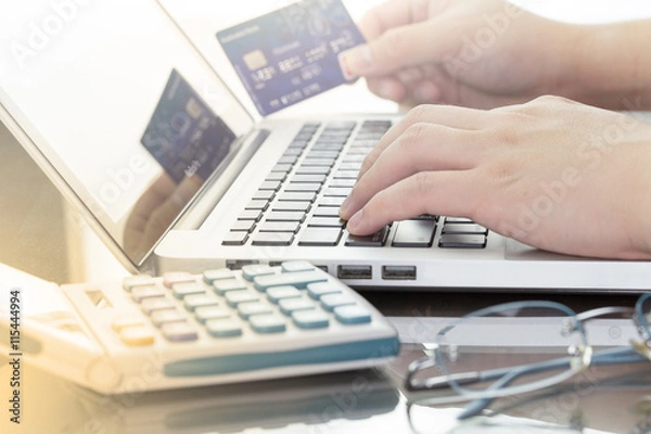Fototapeta Mężczyzna korzystający ze swojego laptopa i posiadający zamiar karty kredytowej na zakupy online, biznes cyfrowy lub e-commerce z kalkulatorem i okularami. ciepły ton.