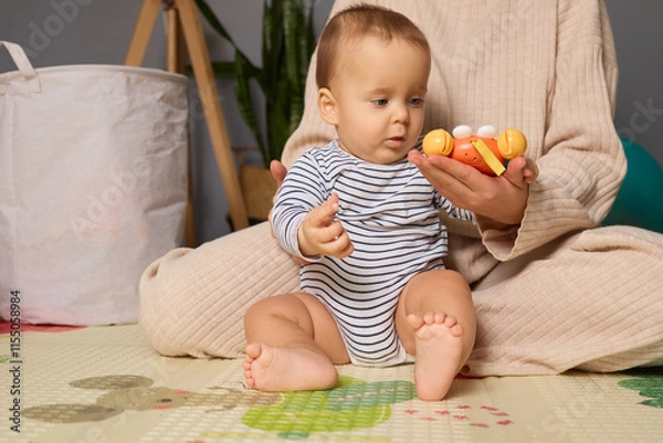 Fototapeta A baby sits comfortably on a play mat, examining a colorful toy held by an adult. The setting is cozy and bright, filled with a warm atmosphere suitable for play
