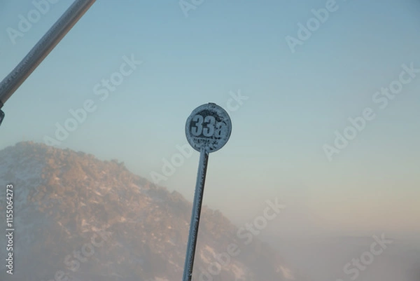 Obraz Close-up of a frosty ski slope marker "33a" against a misty mountain landscape during sunrise.