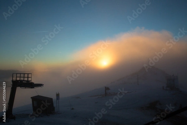 Fototapeta A breathtaking sunrise over a snowy mountain peak, partially covered by fog and mist.