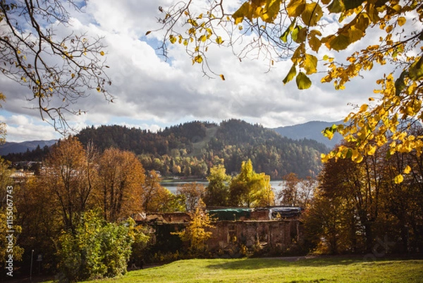 Fototapeta Scenic view of Lake Bled surrounded by vibrant autumn trees and a mountainous landscape under a partly cloudy sky. The golden foliage and rustic charm of the scene capture the beauty of the season