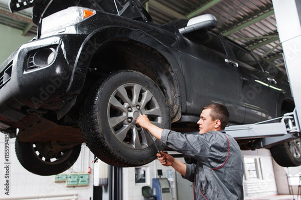Fototapeta Mechanic diagnosing car wheel with screwdriver. Car maintenance in garage. Young auto service worker fixing wheel on raised on hoist car.