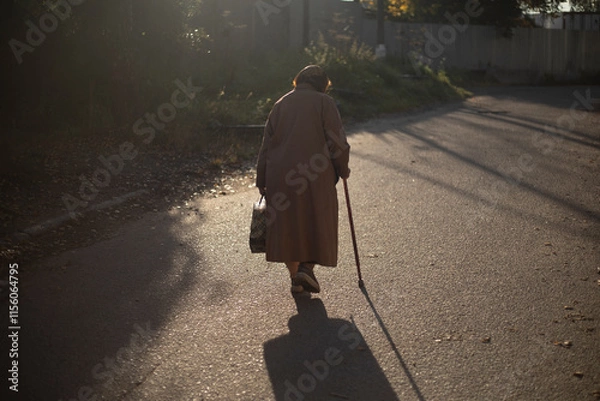 Obraz Senior man walking on road in sunlight. Retired woman with walking stick in autumn. An old woman in a coat.