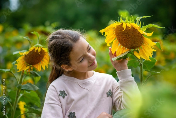 Fototapeta A young girl interacts joyfully with a large sunflower among an array of blooming flowers in a sunlit garden on a pleasant day.
