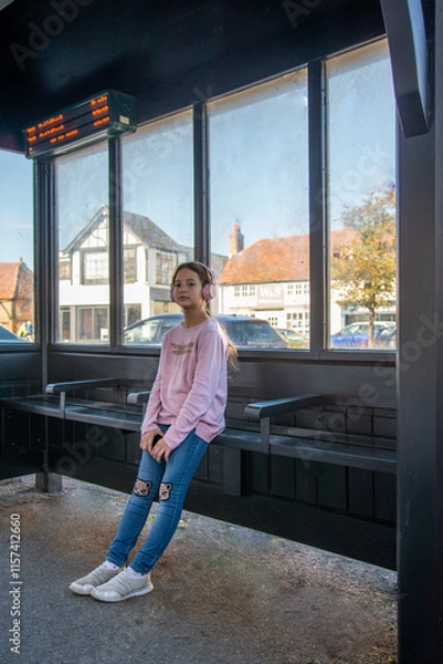 Fototapeta Young girl waiting at a cozy bus stop on a sunny afternoon a charming bus stop, sunlight streaming through large windows, revealing a quiet street and picturesque buildings outside.