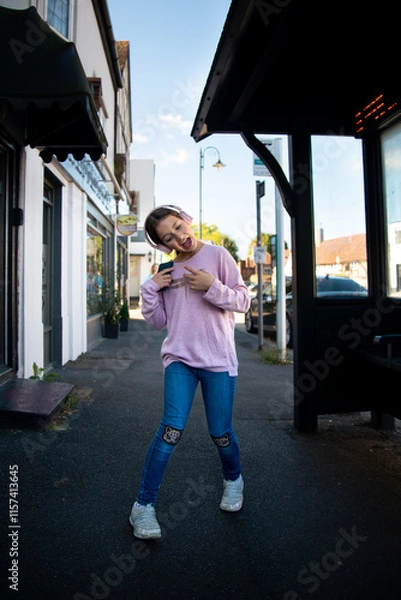 Fototapeta Young girl posing playfully on a vibrant city street during the day.  A girl in a pink sweater joyfully poses with excitement on a lively urban sidewalk, surrounded by charming shops and a clear blue 