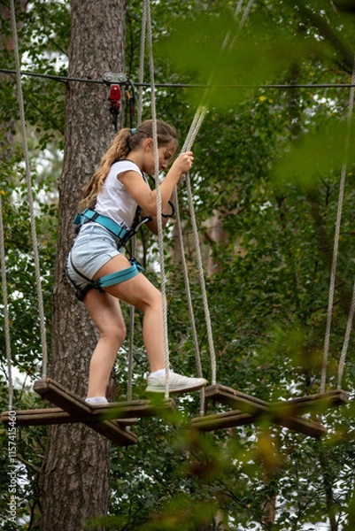 Fototapeta A young girl expertly balances on a swinging bridge high above the ground, surrounded by lush greenery, enjoying an adventurous day in a treetop park.