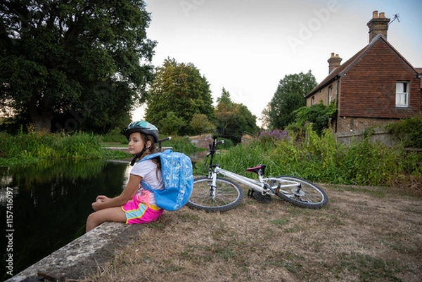 Fototapeta A girl in a helmet takes a break by a calm canal, enjoying nature. Her bicycle lies next to her as she gazes thoughtfully at the serene surroundings.