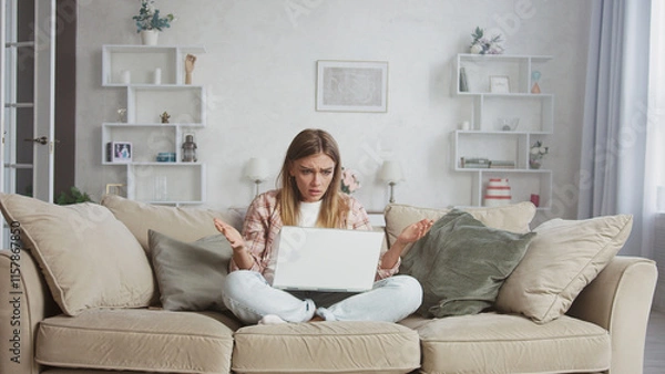 Obraz Young woman sitting with crossed legs on a beige sofa in her living room, looking confused and gesturing in frustration while using a laptop with a blank white screen