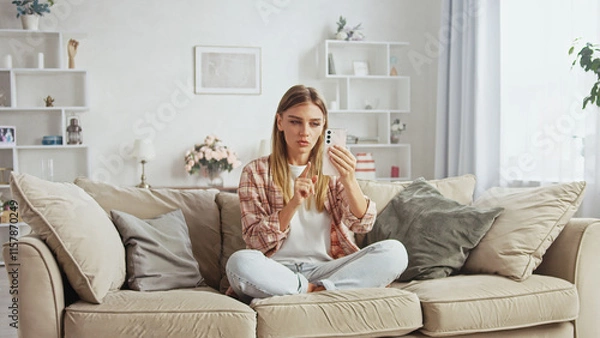 Obraz Young woman sitting with crossed legs on comfortable sofa in cozy living room, making video call using smartphone, pointing finger at screen while talking to someone online