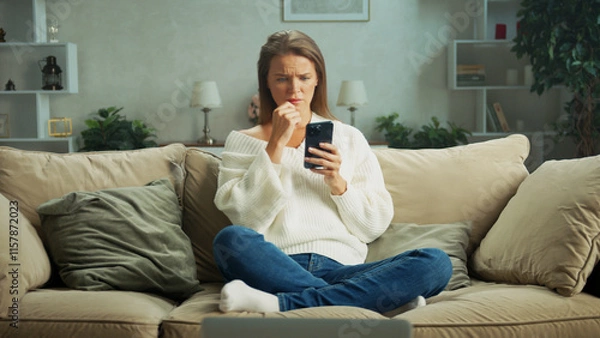 Obraz Young woman sitting on a cozy sofa at home, looking concerned while reading a message on her smartphone and nervously biting her nails, reflecting stress and anxiety