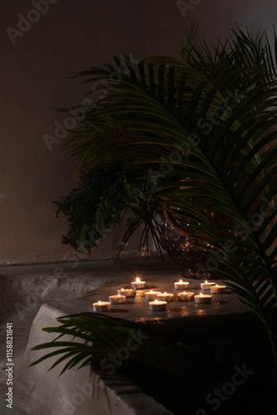 Obraz A tranquil arrangement of tea light candles on a concrete ledge surrounded by lush green palm leaves. The warm glow of the candles contrasts beautifully with the dark, moody background, evoking a sens