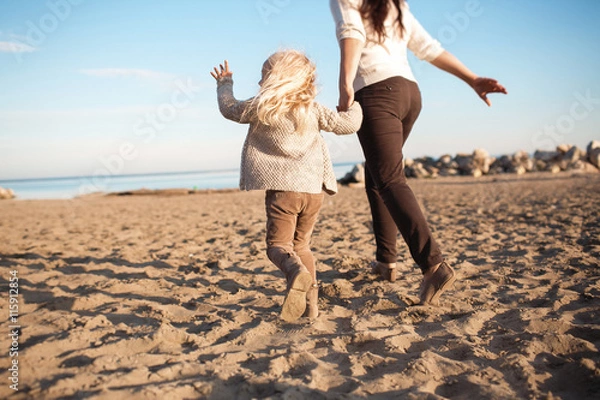 Fototapeta BACK VIEW: Little daughter runs with his mother on a beach