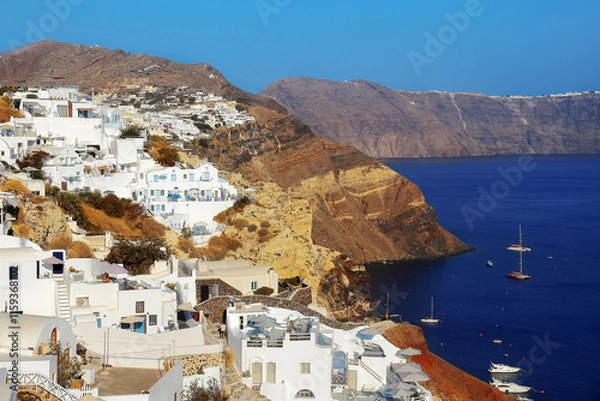 Fototapeta Picturesque view of the traditional white houses on the mountainside in Oia against the backdrop of the sea and Santorini volcano caldera.