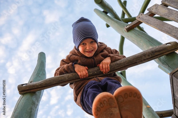 Fototapeta Cheerful child in warm clothing and striped beanie on wooden playground structure with sky in background, low-angle shot. Childhood play and imagination. Health benefits of active lifestyles for kids.