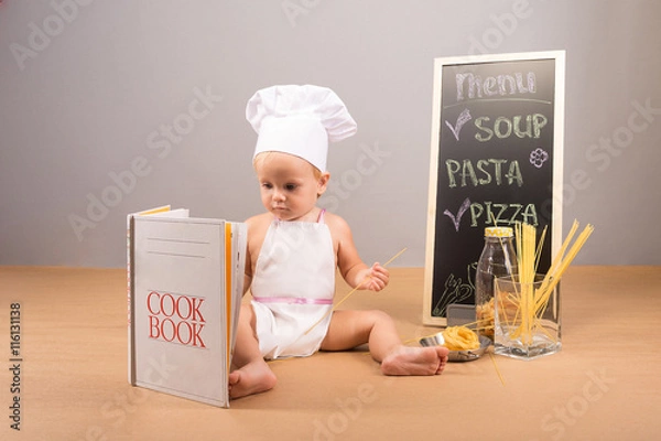 Fototapeta A little boy in a cap and apron cook cookbook studies. Beside him stand menu, spaghetti and colorful pasta
