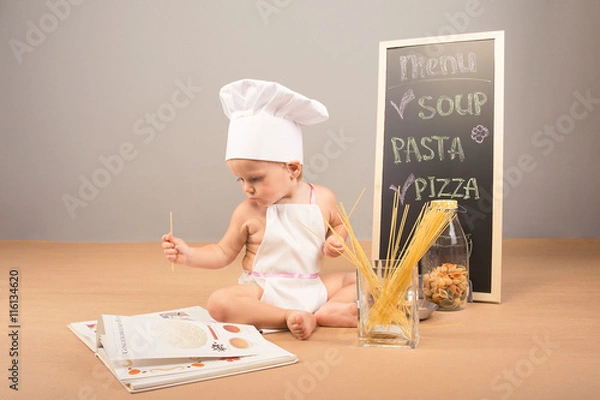 Fototapeta A little boy in a cap and apron cook cookbook studies. Beside him stand menu, spaghetti and colorful pasta