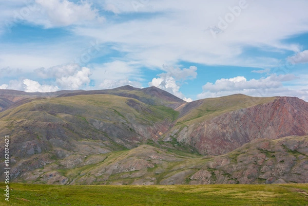 Fototapeta Beautiful layered landscape with multicolor hills and mountains in sunlight under clouds in blue sky. Scenic alpine view to sunlit colorful mountain range of many vivid colors in changeable weather.