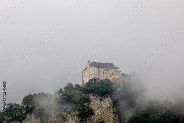 Fototapeta Schloss Tirol in Dorf Tirol near Meran, surrounded by a misty alpine landscape in summer. The historic castle emerges through the fog, creating a mystical and atmospheric scene.