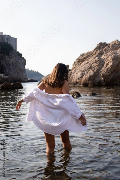 Obraz Girl in white shirt stands in water on the shore of the Adriatic Sea in Croatian Dubrovnik. Young woman in sea water against the backdrop of rocks during summer vacation