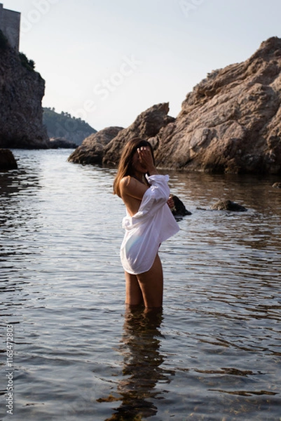 Obraz Girl in white shirt stands in water on the shore of the Adriatic Sea in Croatian Dubrovnik. Young woman in sea water against the backdrop of rocks during summer vacation