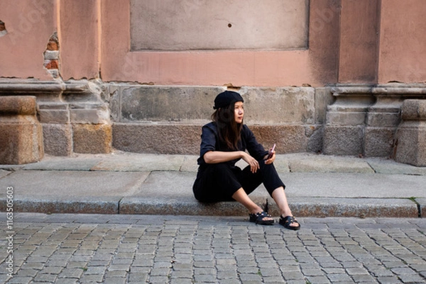 Obraz Girl sitting on the pavement in the city. Young woman in total black style and a docker cap