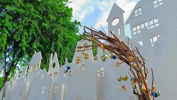 Fototapeta A charming artistic display showcasing a whimsical arch made of twigs, embellished with colorful ornaments, set against a backdrop of white, stylized houses and vibrant greenery