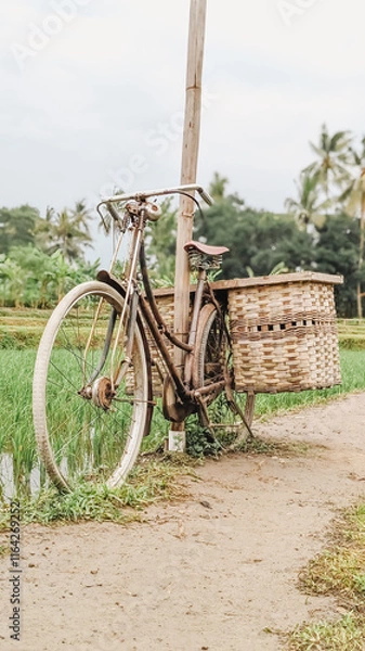 Obraz A classic bicycle with bambo-made baskets leaning against a bamboo post beside scenic rural paddy fields conveying nostalgia simplicity and rustic charm ideal for rural travel and lifestyle themes