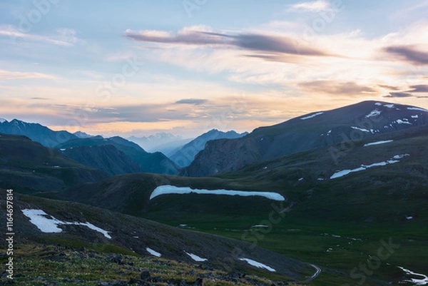 Fototapeta Scenic view from flowering grassy stony hillside to alpine valley, green pass, deep gorge and big sharp mountain peak silhouette against large snowy top far away under vivid sunset color cloudy sky.