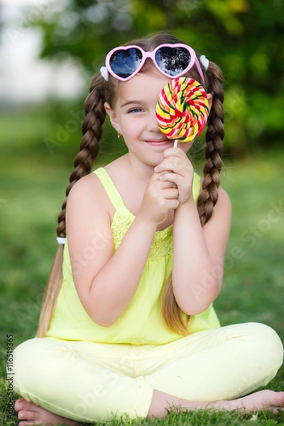 Fototapeta The girl with pigtails,wearing sunglasses in pink frame with glass in the form of hearts,dressed in a yellow t-shirt and pants sitting on green grass in summer Park with a large round candy on a stick