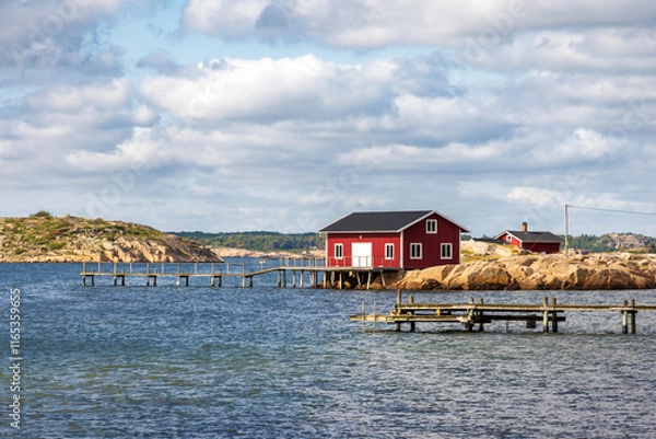 Fototapeta Seascape on the Resö island in the Bohuslän province, Sweden, showing a typical red painted boathouse on the rocky coast with a wooden pier under a stormy sky on a sunny summer day.