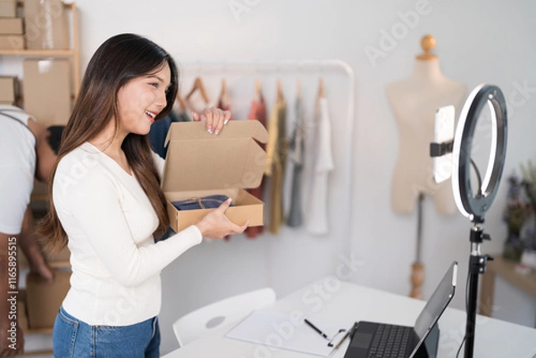 Obraz  A woman enthusiastically livestreams while holding a box, showcasing her products with a ring light and laptop setup in a workspace filled with racks and packaging