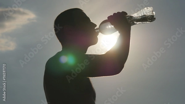 Obraz Young man drinking water from a plastic bottle in nature. Guy having water break at sunset. The Sun in the Background. Silhouette of male profile