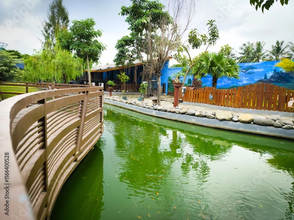 Fototapeta Beautiful Wooden Bridge Over Green Pond With Serene Environment and Lush Green Trees