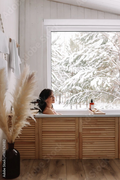 Obraz A young woman is relaxing lying in the bathtub in a cozy bathroom with panoramic windows overlooking a snowy forest in a country house in winter. Girl takes water treatments at a spa. Selective focus 