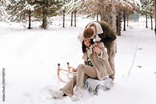 Fototapeta A man and a woman in love ride a sleigh through a snowy forest in winter, relaxing in the countryside during the Christmas holidays. Selective focus