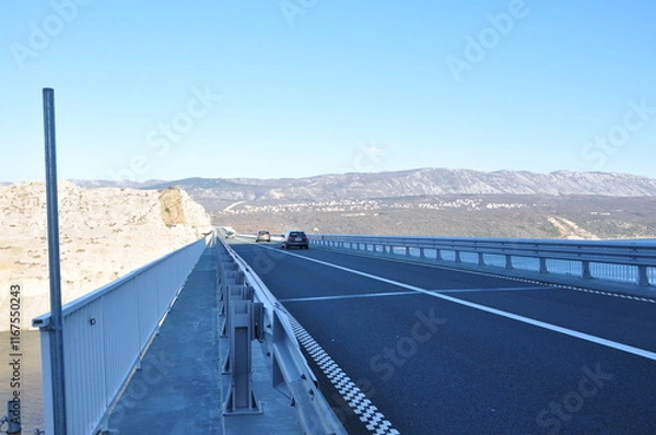 Fototapeta Arched concrete bridge spanning across a tranquil body of water, on the backdrop of green hills and distant mountains. Krk bridge, Croatia