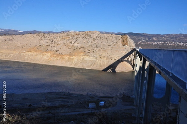 Fototapeta Arched concrete bridge spanning across a tranquil body of water, on the backdrop of green hills and distant mountains. Krk bridge, Croatia