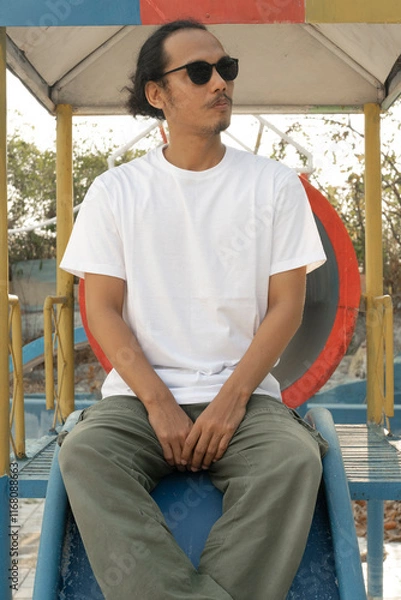Fototapeta An Asian male model wearing a plain white t-shirt and black sunglasses is posing in a commercial park during the daytime. The male model's white t-shirt is being used as a mockup object for showcasing